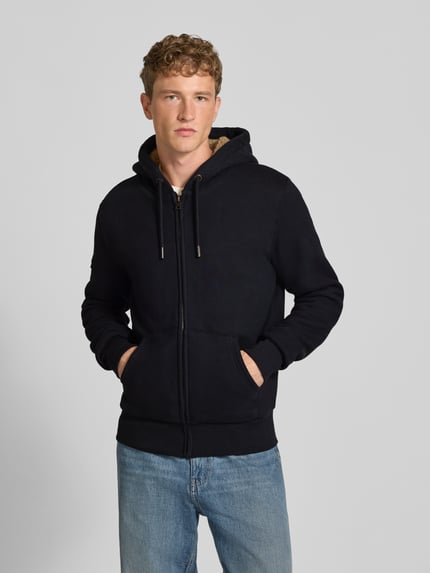 Mężczyzna nosi Regular Fit Sweatjacke mit geteilter Känguru-Tasche Modell 'Borg' von Superdry, czarny kolor, kaptur z tunelzugiem, kieszeń kangurka, długość tyłu w rozmiarze M: 71 cm, długość rękawów w rozmiarze M: 72 cm, widok całej sylwetki z przodu