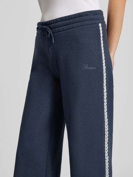 Frauenmodel trägt Wide Fit Sweatpants aus Baumwoll-Mix von REVIEW in Dunkelblau mit Bordüre und Label-Stitching, Nahaufnahme Oberkörper.