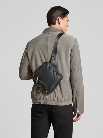 Männermodel trägt Crossbody Bag mit Statement-Print Modell 'EXPLORE' von camel active, schwarze Farbe, Reißverschlussfächer, Ganzkörperansicht Rückansicht.