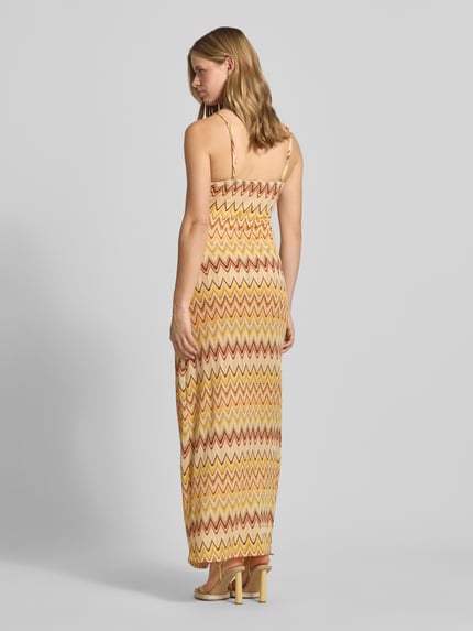 Vrouwenmodel draagt Maxikleid im Allover-Look van Gina Tricot in materiaalmix met stretch, recht model, spaghettibandjes, ruglengte maat S 150 cm, borstwijdte 32 cm, all-over look in geel en oranje tinten, detail van zigzagpatroon, rugaanzicht full-body.