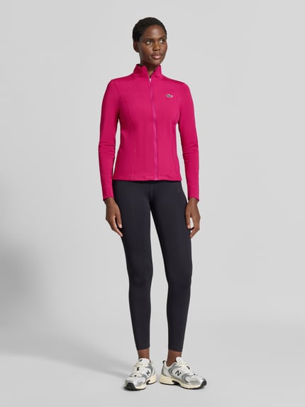Frauenmodel trägt Slim fit Trainingsjacke mit Logo-Patch von Lacoste in Pink und Schwarz, glatte Struktur, Damen Trainingsjacke, Ganzkörperansicht von vorne.