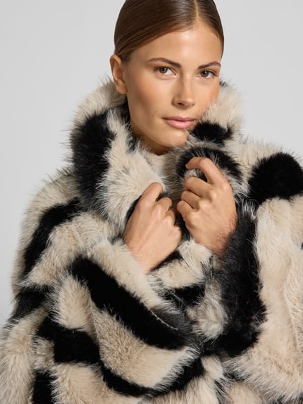 Frauenmodel trägt Mantel aus Kunstfell im Allover-Look von Beaumont Amsterdam, beige und schwarz, weiche Textur, Nahaufnahme Oberkörper, Relaxed Fit, Reverskragen, Knopfverschluss, KI-modifiziertes Bild.