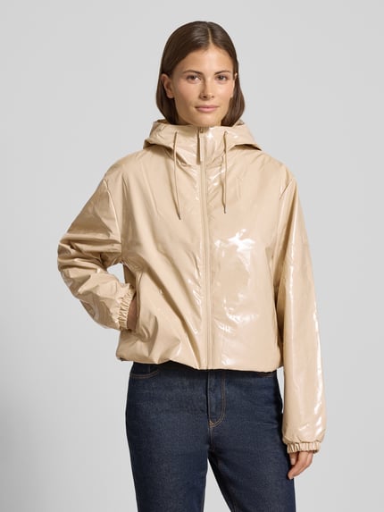 Frauenmodel trägt Jacke mit Kapuze Modell 'Lohja' von RAINS in Beige mit Reißverschluss, lockerem Schnitt, Label-Detail, Ganzkörperansicht von vorne.