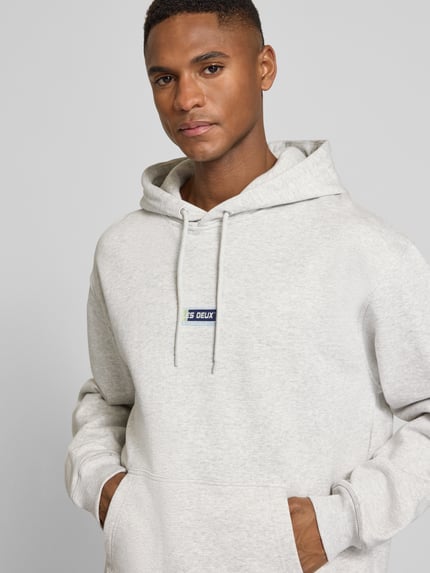 Männermodel trägt Hoodie mit Logo-Print Modell 'Chase' von Les Deux aus Reine Baumwolle im Loose-Fit mit Kapuze, Kordelzug, Känguru-Tasche, elastische Abschlüsse, hellgraue Farbe, melierte Textur, Nahaufnahme Oberkörper.