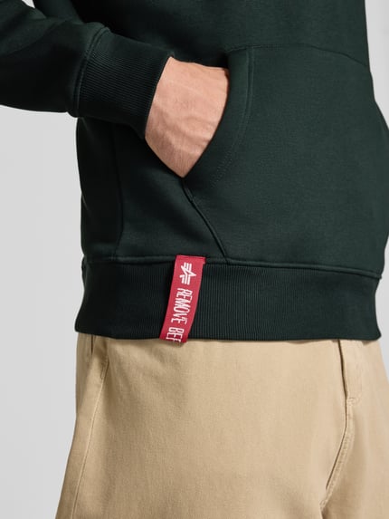Männermodel trägt Regular Fit Hoodie aus Baumwoll-Mix mit Logo-Print von Alpha Industries, dunkelgrün, beige Hose, Nahaufnahme Oberkörper, Label am Saum sichtbar.