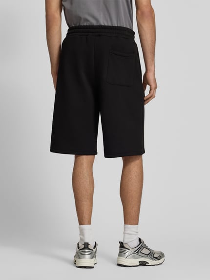 Männermodel trägt Sweatshorts mit Logo-Print von REVIEW in Schwarz, Material-Mix mit Baumwoll-Anteil, Loose-Fit, elastischer Bund, Ganzkörperansicht Rückansicht.