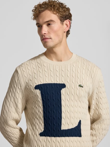 Männermodel trägt Regular Fit Strickpullover aus Baumwoll-Mix von Lacoste in Creme mit blauem L und Logo, Nahaufnahme Oberkörper. Herren Strickjacke von Lacoste.