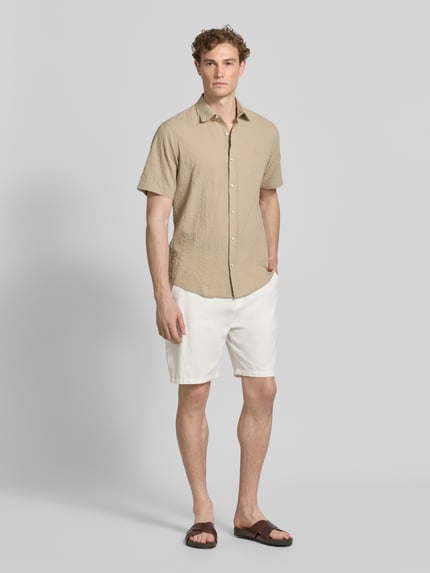 Männermodel trägt Relaxed Fit vrijetijdsoverhemd met logostitching en kentkraag van Lerros, seersucker, knoopsluiting, afgeronde zoom, ruglengte bij maat M 75 cm, schouderbreedte 45 cm, mouwlengte 25 cm, zandkleurig, witte korte broek, bruine sandalen, ganzkörperansicht van voren.