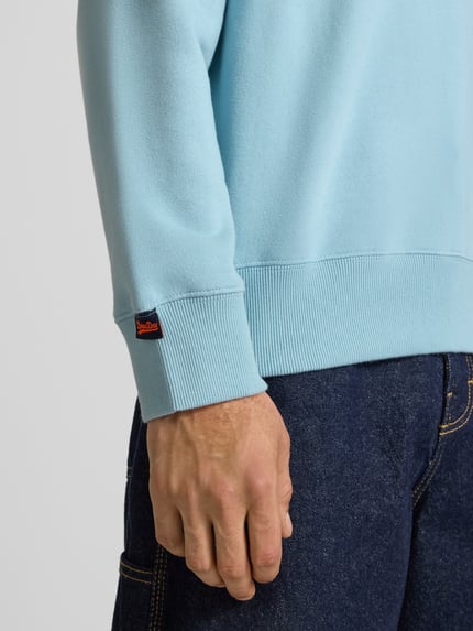 Männermodel trägt Sweatshirt mit Rundhalsausschnitt und Label-Stitching von Superdry in Hellblau, Baumwoll-Mix, Nahaufnahme Oberkörper. Regular-Fit, Rückenlänge bei Größe M 61 cm, Ärmellänge 64 cm.