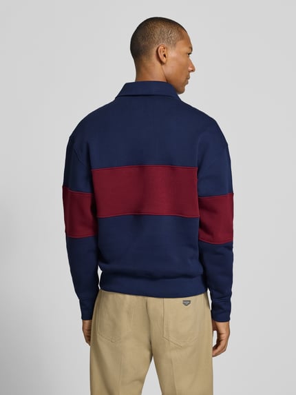 Männermodel trägt Relaxed Fit Sweatshirt aus reiner Baumwolle von ARMEDANGELS in Dunkelblau mit bordeauxfarbenem Streifen, strukturierte Oberfläche, Ganzkörperansicht Rückansicht.