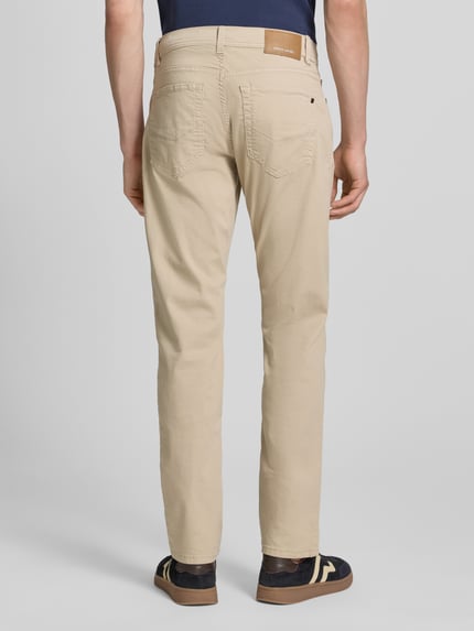 Męski model nosi Jeans mit Label-Detail Modell 'Lyon' von Pierre Cardin, bawełniany mix ze stretchem, krój regular fit, beżowy kolor, widoczne kieszenie i detal z logo, Szerokość w rozmiarze 32/32: 41 cm, długość nogawki wewnętrznej: 80 cm, widok całej sylwetki z tyłu.