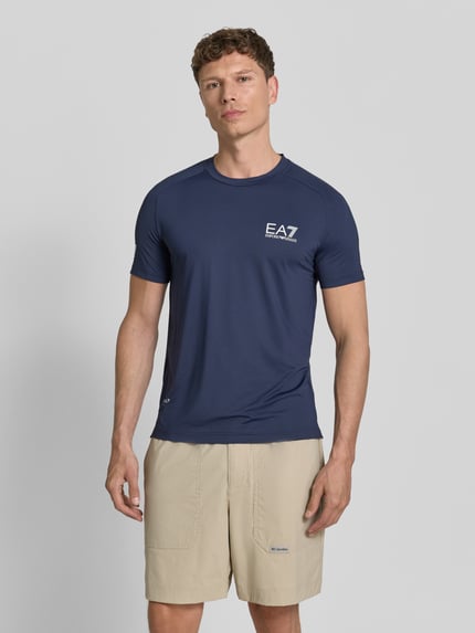 Męski model nosi T-Shirt mit Label-Print od EA7 Emporio Armani, materiał z domieszką stretchu, krój slim fit, granatowy kolor, całkowity widok z przodu.