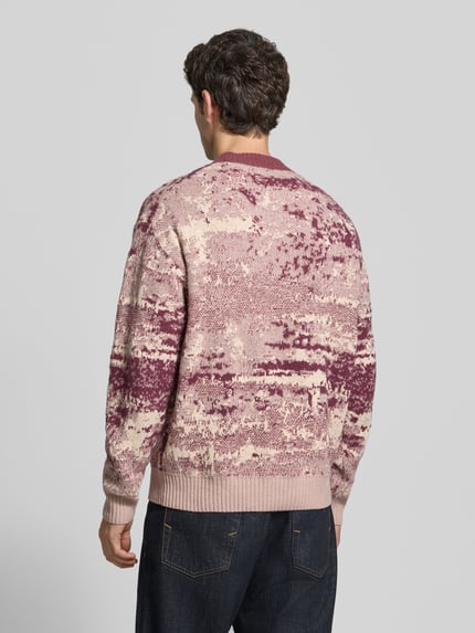 Männermodel trägt Pullover mit geripptem Rundhals Modell 'Kastet' von BOSS Orange, mehrfarbiges Strickmuster, Ganzkörperansicht Rückansicht.
