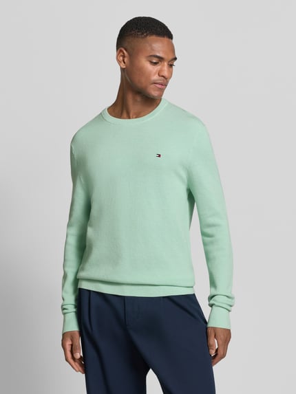 Männermodel trägt Regular Fit Strickpullover aus reiner Baumwolle von Tommy Hilfiger in Optik: Unifarben, Strickoptik, Logo-Detail, Ganzkörperansicht von vorne.