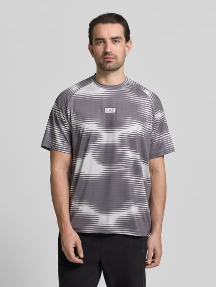 Mężczyznamodel nosi Regular Fit T-Shirt mit Logo-Applikation od EA7 Emporio Armani, elastyczny materiał, krój regular fit, okrągły dekolt, szaro-białe wzory, długość tyłu w rozmiarze M: 73 cm, widok całej sylwetki z przodu.