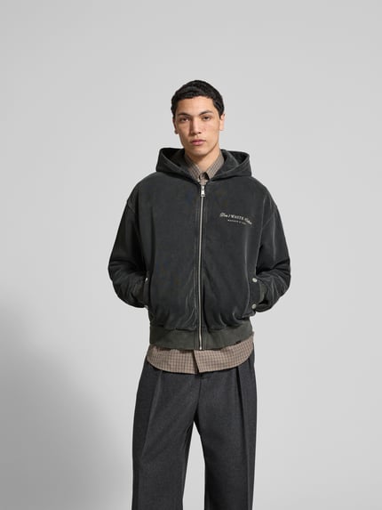 Männermodel trägt Oversized Jeansjacke aus Baumwoll-Mix von Don't Waste Culture, dunkle Farbe, Eingrifftaschen, Ganzkörperansicht von vorne. Rückenlänge bei Größe M 65 cm, Schulterbreite 58 cm, Ärmellänge 67 cm, durchgehender Reißverschluss, elastische Abschlüsse, Druckknopfverschluss an den Taschen.