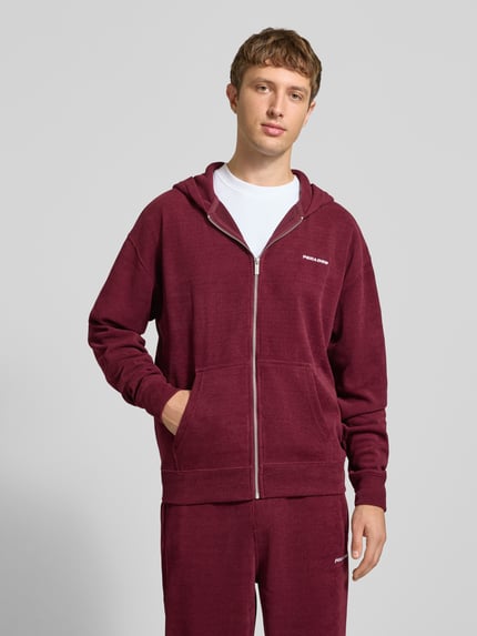 Männermodel trägt Oversized Fit Sweatjacke mit Label-Detail Modell 'Mezzano' von PEGADOR in Dunkelrot, Baumwoll-Material-Mix, getrennte Känguru-Tasche, elastische Abschlüsse, Ganzkörperansicht von vorne.