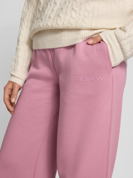 Frauenmodel trägt Straight Fit Sweatpants aus Baumwoll-Mix mit Label-Detail von REVIEW in Rosa, strukturierter Stoff, Nahaufnahme Oberkörper. Damen Sweatpants von REVIEW, Baumwoll-Mix, Straight Fit, Label-Detail, elastischer Bund, Innenbeinlänge bei Größe S 79 cm.