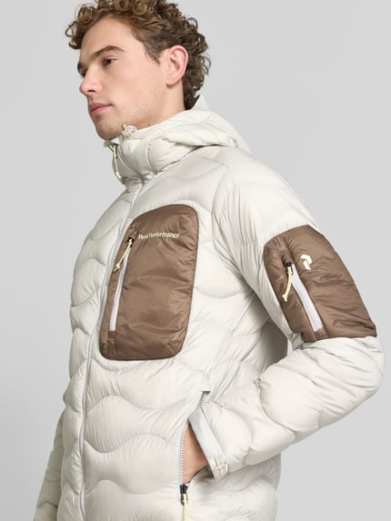 Męski model nosi kurtka puchowa z kontrastującą brustaszą Daunenjacke mit kontrastierender Brusttasche von Peak Performance, dwukolorowy, matowy, faktura, krój regular fit, długi, aplikacja z logo, zamki błyskawiczne, kaptur, pasek do ściągania, kieszenie zapinane na zamek błyskawiczny, kieszeń boczna naszywana, kieszeń zapinana na zamek błyskawiczny, kaptur, Ganzkörperansicht z przodu.