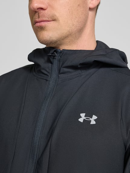 Mężczyznamodel nosi Regular Fit Jacke mit Logo-Print Modell 'Unstoppable' von Under Armour, elastyczna mieszanka materiałów, krój regular fit, nadruk z logo, wpuszczane kieszenie zapinane na zamek błyskawiczny, zamek błyskawiczny, kaptur ze stójką, długość tyłu w rozmiarze M: 74 cm, długość rękawów w rozmiarze M: 68 cm, ciemny kolor, gładka tekstura, zbliżenie na górną część tułowia.