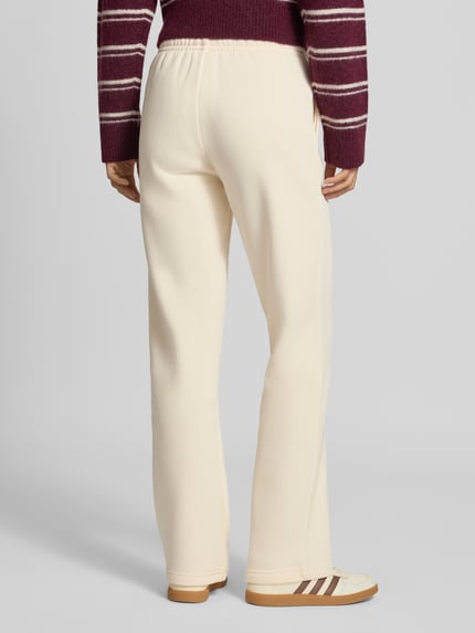 Männermodel trägt Wide Fit Sweatpants aus Baumwoll-Mix Modell 'Iconic' von Gina Tricot, cremefarben, glatte Textur, elastischer Bund, Ganzkörperansicht Rückansicht, kontekst: damskie spodnie dresowe, bawełniany miks, szeroki krój, elastyczny pas ze sznurkiem, długość nogawki w rozmiarze S: 80 cm.