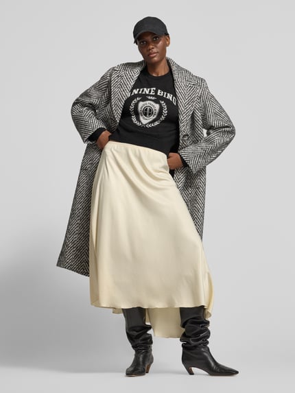 Frauenmodel trägt Sweatshirt mit Logo-Front Modell 'MIA SWEATER' von Anine Bing in Melange-Optik, Motiv-Print, überschnittene Schultern, Rundhals; dazu heller Maxirock, gemusterter Mantel, schwarze Stiefel, Ganzkörperansicht von vorne.