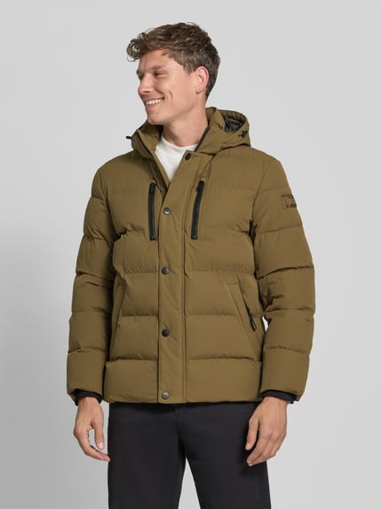 Männermodel trägt Steppjacke mit Kapuze von BRAX in Unifarbe, schwarze Reißverschlüsse, Logo-Patch, Ganzkörperansicht von vorne.