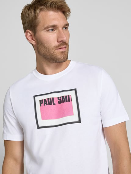 Männermodel trägt T-Shirt mit Label-Print von Paul Smith in Weiß mit rosa und schwarzem Print, reine Baumwolle, Regular Fit, Rundhalsausschnitt, Nahaufnahme Oberkörper.