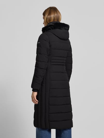 Frauenmodel trägt schwarzen Steppmantel mit Kunstfellkapuze und Zweiwege-Reißverschluss von NAUTGÅRD 'ECLIPSE', Ganzkörperansicht Rückansicht. Rückenlänge 115 cm, Schulterbreite 40 cm, Brustweite 46 cm, Ärmellänge 65 cm, Slim Fit, glatte Struktur.