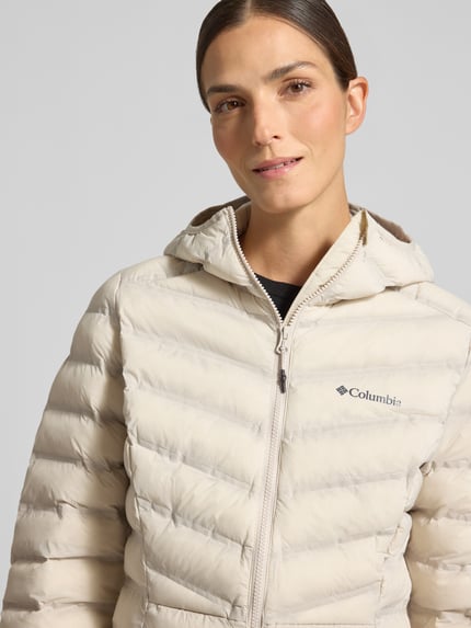 Frauenmodel trägt Jacke in Stepp-Optik Modell 'Terwilliger' von Columbia in hellem Beige mit Logo-Print, Nahaufnahme Oberkörper.