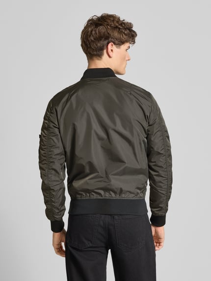 Męski model ma na sobie Regular Fit Bomberjacke mit Logo-Patch od Alpha Industries, ciemnozielony materiał o gładkiej fakturze, widok całej sylwetki z tyłu, długość tyłu 65 cm, szerokość ramion 45 cm, szerokość w rozmiarze M 54 cm, długość rękawa 69 cm