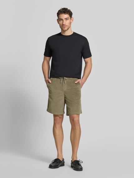 Męski model nosi Regular Fit Sweatshorts aus Baumwoll-Mix von SELECTED HOMME, jasnooliwkowe, sztruks, krój regular fit, kieszenie boczne i tylna, elastyczny pas ze sznurkiem, widok całej sylwetki z przodu.