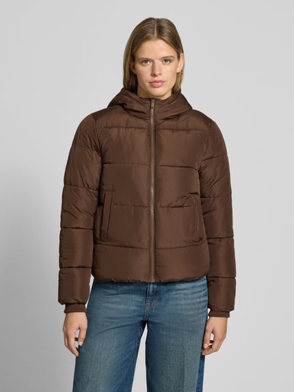 Frauenmodel trägt braune Steppjacke mit Kapuze von PIECES zur blauen Jeans, Matte Optik, Frontreißverschluss, Ganzkörperansicht von vorne.