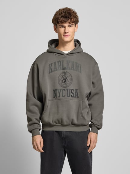 Männermodel trägt Hoodie mit Label-Print und Känguru-Tasche von KARL KANI aus Baumwoll-Mix in Grau, oversized, Ganzkörperansicht von vorne.