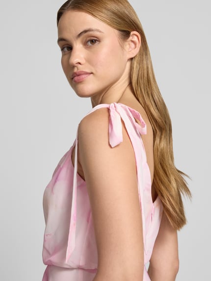 Vrouwenmodel draagt knielanges cocktailjurk met bloemen all-over print van Jake*s Cocktail, sierstrik, mouwloos design, gevoerde onderrok, getailleerde pasvorm, pastelroze tinten, detailopname bovenlichaam.