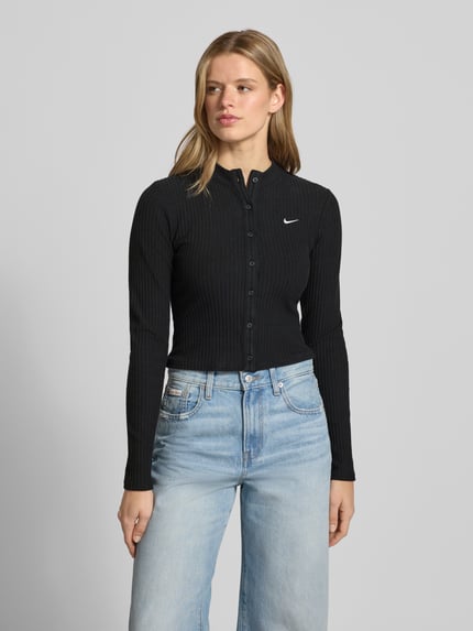 Frauenmodel trägt Cardigan in Ripp-Optik Modell 'CHILL' von Nike, Skinny-Fit, Stehkragen, Label-Stitching, Langärmelig, Durchgehende Knopfleiste, schwarze Farbe mit feiner Ripp-Struktur, Jeans kombiniert, Nahaufnahme Oberkörper