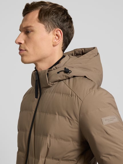 Mężczyzna model nosi Regular Fit kurtka puchowa z kapturem od Marc O'Polo, jednokolorowa, zamek błyskawiczny dwustronny, funkcje: wodoszczelna, wiatroszczelna, oddychająca, kieszenie boczne i wewnętrzne, długi rękaw elastyczne lamówki, wypełnienie z puchu i pierza; zbliżenie na górę ciała z przodu.