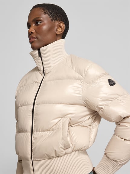 Frauenmodel trägt Funktionsjacke mit Logo-Patch Modell 'queens bomber' von Moose Knuckles in Beige, Steppstruktur, gerippte Bündchen, Nahaufnahme Oberkörper.