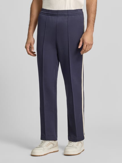 Männermodel trägt Regular Fit Jogpants aus Baumwoll-Mix von Lacoste in Dunkelblau mit beigen Seitenstreifen, Herren Weite Hose, Ganzkörperansicht von vorne.