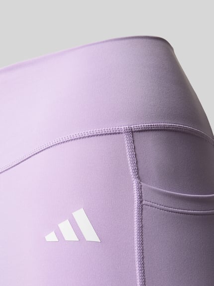 Produktfoto, fioletowe Slim Fit Leggins mit Logo-Print von adidas Sportswear, elastyczny materiał, szeroki pas, zbliżenie na biodro