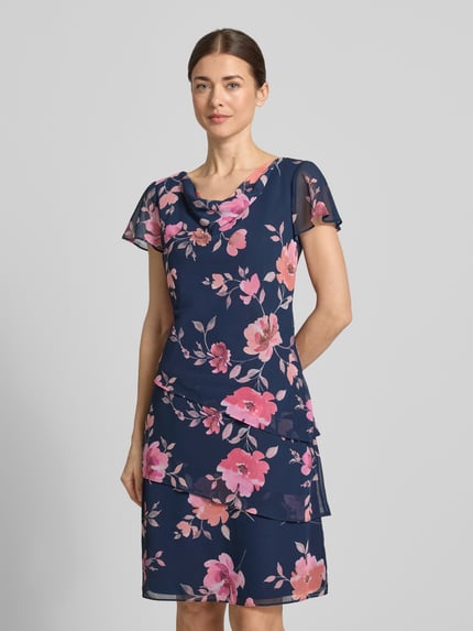 Modelka kobieca nosi Knielanges Cocktailkleid mit Wasserfall-Ausschnitt od Christian Berg Cocktail, granatowa tkanina szyfonowa z różowymi kwiatami, krój taliowany, długość do kolan, dekolt woda, rękawy motylkowe, spódnica z drapowaniami, długość tyłu w rozmiarze 36: 97 cm, szerokość w rozmiarze 36: 46 cm, długość rękawa w rozmiarze 36: 16 cm, widok całej sylwetki z przodu.
