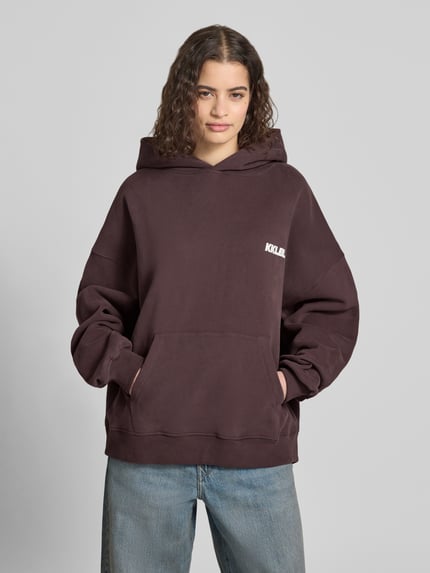 Frauenmodel trägt Oversized Hoodie mit Label-Prints und Känguru-Tasche von Karo Kauer, braune Baumwolle, lockere Form, Ganzkörperansicht von vorne.