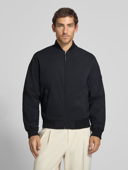Męski model nosi bomberkę z zamkiem błyskawicznym i wpuszczanymi kieszeniami Bomberjacke mit Reißverschluss und Eingrifftaschen von JACK & JONES, krój regular fit, stójka, długość tyłu w rozmiarze M: 66 cm, szerokość ramion w rozmiarze M: 42 cm, szerokość klatki piersiowej w rozmiarze M: 60 cm, długość rękawów w rozmiarze M: 68 cm, czarny kolor, gładka tekstura, całkowity widok z przodu.