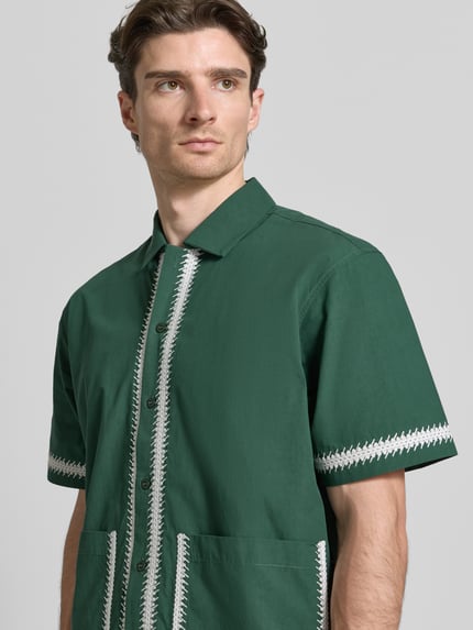 Männermodel trägt Regular Fit Freizeithemd mit aufgesetzten Taschen von WOODBIRD in grün mit Kentkraag, Stichdetails, Nahaufnahme Oberkörper.