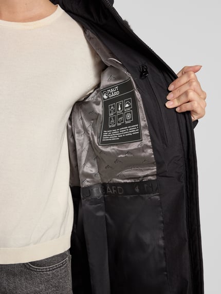 Frauenmodel trägt Jacke mit abnehmbarer Kapuze Modell 'ECLIPSE' von NAUTGÅRD, schwarze Außenseite, silbergraues Innenfutter mit Label-Patch, Nahaufnahme Oberkörper, Kontext: gerader Schnitt, Zweiwege-Reißverschluss, seitliche Eingrifftaschen mit Druckknopfverschluss, gestrickte Ärmelabschlüsse, Rückenlänge 79 cm, Schulterbreite 38 cm, Brustweite 48 cm, Ärmellänge 62 cm.