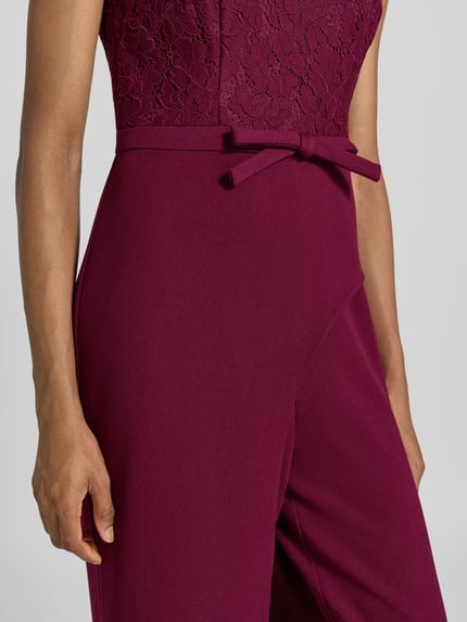 Kobieta nosi Regular Fit Jumpsuit mit Zierschleife od Adrianna Papell, mieszanka materiałów z dodatkiem wiskozy, ozdobna kokarda, obszycie koronką, kwiatowy wzór, krótkie rękawy, głęboki burgund, tekstura gładka i koronkowa, szerokość w rozmiarze 36: 40 cm, długość tyłu: 37 cm, całkowita długość: 150 cm, długość wewnętrzna nogawki: 84 cm, zbliżenie na górną część kombinezonu