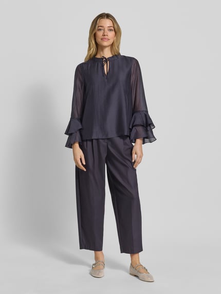 Frauenmodel trägt Regular Fit Bluse aus Lyocell-Mix mit Volants von Comma in dunklem Blau, Schlüsselloch-Ausschnitt, fließende Struktur, Rückenlänge bei Größe 36: 58 cm, Ärmellänge 60 cm, Ganzkörperansicht von vorne.