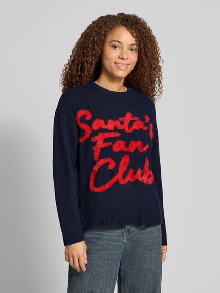 Frauenmodel trägt Pullover mit Santa Schriftzug von ONLY in mehrfarbiger Strickoptik mit Motiv-Print, Rundhals, Ganzkörperansicht von vorne.