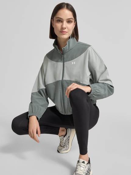 Modelka kobieca nosi kurtkę treningową z stójką i wpuszczanymi kieszeniami Under Armour, krój regular fit, logo, zamek błyskawiczny, długość tyłu w rozmiarze S: 60 cm, szerokość ramion w rozmiarze S: 62 cm, szerokość klatki piersiowej w rozmiarze S: 57 cm, długość rękawów w rozmiarze S: 50 cm, biało-szaro-zielone barwy, geometryczne panele, gładka tekstura; całkowity widok z przodu.