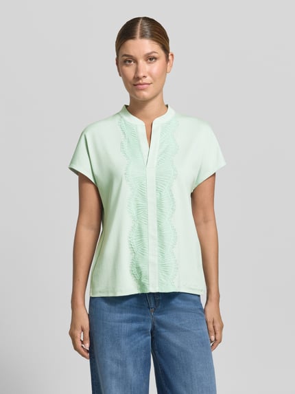 Kobiece model prezentuje Regular Fit Bluse aus Baumwoll-Mix mit Spitzenbesatz od Marc Cain w kolorze jasnej mięty, mieszanka bawełny, krój regular fit, obszycie koronką, dekolt serafino, z krótkim rękawem, długość tyłu w rozmiarze 36: 57 cm, zdjęcie całej sylwetki z przodu.